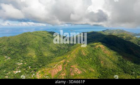Üppige, grüne Hügel kaskadieren in Richtung Ozean unter einem Himmel von Wolken, mit kleinen Inseln am Horizont. Praslin, Seychellen. Stockfoto