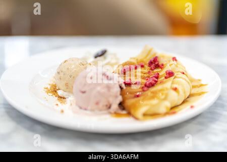 Dessertteller mit weichem Fokus und gefalteten Crepes, mit Sirup getränkt und mit Himbeeren belegt, serviert mit zwei Kugeln Eis Stockfoto
