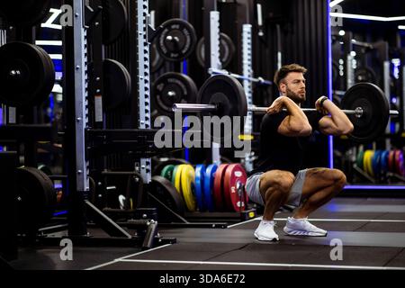 Ein Mann konzentriert sich intensiv, während er eine Langhantelhocke in einem gut ausgestatteten Fitnessstudio macht. Die kräftigen Gewichte auf den Racks verleihen dem Hintergrund Farbe, Showcasin Stockfoto