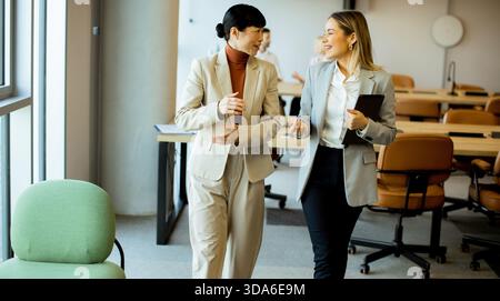 Zwei Frauen in Geschäftskleidung gehen und reden in einem hellen, modernen Büro. Sie scheinen engagiert und freundlich zu sein und teilen Ideen in einem kollaborativen Umfeld Stockfoto