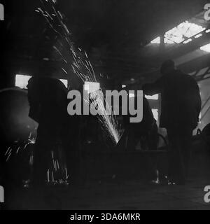 Slawjansk, Ukrainische SSR, UdSSR - 1970er Jahre: Sowjetische Metallarbeiter schleifen schwere Maschinenteile in der Werkstatt der Stroymash-Fabrik. Die Silhouette eines Mannes, der mit einer Schleiferei arbeitet und einen Schauer von hellen Funken vor dem Hintergrund großer Industriefenster erzeugt. Dieses stimmungsvolle Schwarz-weiß-Archivfoto zeigt den intensiven Herstellungsprozess und die harte Handarbeit in der friedlichen Donbass-Region während der Sowjetzeit. Metallurgie und Produktionsszene der Schwerindustrie. Stockfoto