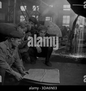 Slawjansk, Ukrainische SSR, UdSSR - 1970er Jahre: Eine Arbeiterbrigade, die einen technischen Plan auf dem Boden der Stroymash-Fabrikwerkstatt untersucht. Das Team von Männern in flachen Kappen und Arbeitsjacken diskutiert sorgfältig das Engineering-Konzept für die Produktion schwerer Maschinen. Dieses stimmungsvolle Schwarz-weiß-Archivfoto fängt den ernsten Moment der industriellen Planung und Kollektivarbeit in der friedlichen Donbass-Region während der Sowjetzeit ein. Teamarbeit in einer Schwerindustrie. Stockfoto
