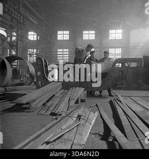 Slawjansk, Ukrainische SSR, UdSSR - 1970er Jahre: Eine Gruppe sowjetischer Ingenieure und Arbeiter diskutierte einen technischen Plan, während sie auf einem massiven Metallrahmen in der Werkstätte Stroymash stand. Das Team besteht aus einem Vorarbeiter in einem Hemd und zwei Arbeitern in flachen Kappen, die die Abmessungen eines Fahrgestells für schwere Maschinen anhand der Zeichnung überprüfen. Dieses Schwarzweiß-Archivfoto fängt die Atmosphäre der industriellen Planung, Teamarbeit und technischen Präzision in der friedlichen Donbass-Region während der Sowjetzeit ein. Stockfoto