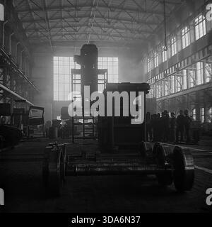 Slawjansk, Ukrainische SSR, UdSSR - 1970er Jahre: Atmosphärischer Innenblick der Werkshalle Stroymash mit einer riesigen mobilen Industrieeinheit im Bau. Schwere Schienenräder auf einer Achse liegen im Vordergrund, während eine Gruppe von Arbeitern im Abstand vor großen Fenstern Silhouetten trägt. Dieses Schwarzweiß-Archivfoto zeigt die kolossale Größe der Schwermaschinenproduktion und die industrielle Macht der friedlichen Donbass-Region während der Sowjetzeit. Produktionsanlage. Stockfoto