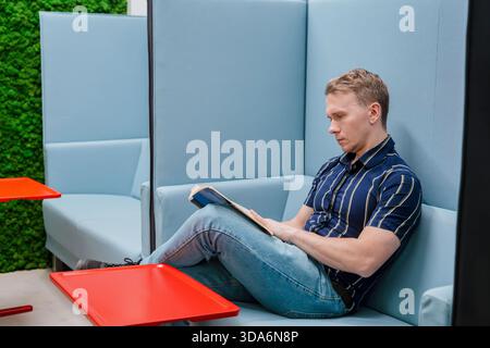 Ein junger Mann liest ein Buch, während er auf der Couch sitzt Stockfoto