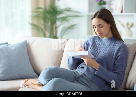 Frau in blauer Schrift Adresse im Umschlag, die zu Hause auf einem Sofa sitzt Stockfoto