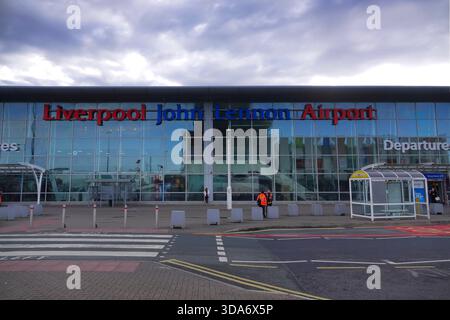 Frontansicht auf Liverpool John Lennon Airport Terminal mit großer Glasfassade, Schildern, Zebraüberquerung und Menschen draußen unter bewölktem Himmel. Stockfoto