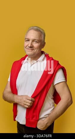 Senior Mann lächelt selbstbewusst auf gelbem Hintergrund in weißem Polo und rotem Pullover Stockfoto