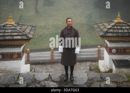 Blick auf einen Mann in traditioneller bhutanischer Kleidung, der selbstbewusst auf Steintreppen steht, flankiert von verzierten Chorden, vor nebeliger Kulisse, Hungtso, Bhutan Stockfoto