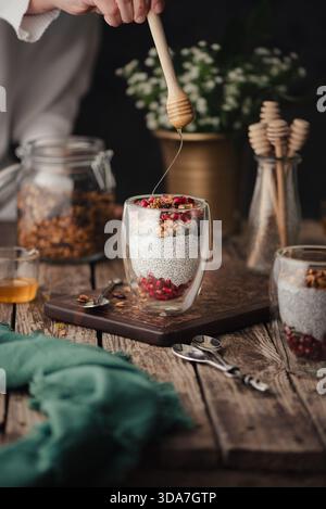 Ein filmisches Lebensmittelfoto, das Chia Pudding mit Joghurt, Granatapfelkernen und Müsli zeigt, während eine Hand Honig aus einem Holzlöffel gießt. S Stockfoto