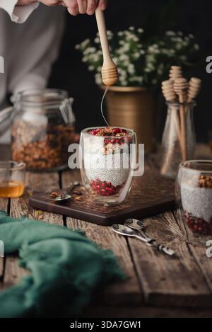 Ein warmes und einladendes Lebensmittelfoto mit Chia-Puddinggläsern mit Granola- und Granatapfelkernen, mit einer Hand, die einen Honiglöffel über dem hält Stockfoto