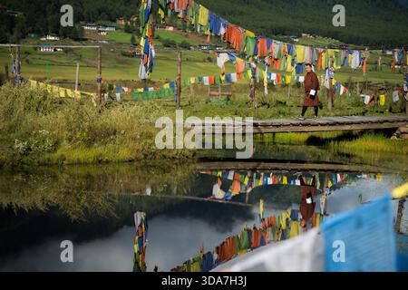 Blick auf Gebetsfahnen, die über eine hölzerne Brücke über einem ruhigen Teich, der den Himmel reflektiert, mit einer Person, die vorbeigeht, Nubding, Wangdue Phodrang, Bhutan. Stockfoto