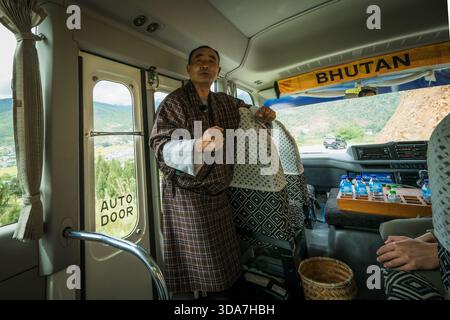 Blick auf einen Reiseleiter in traditioneller bhutanischer Kleidung steht in einem Bus und bewegt sich in Richtung der Landschaft, die durch das Fenster sichtbar ist, Paro, Bhutan. Stockfoto