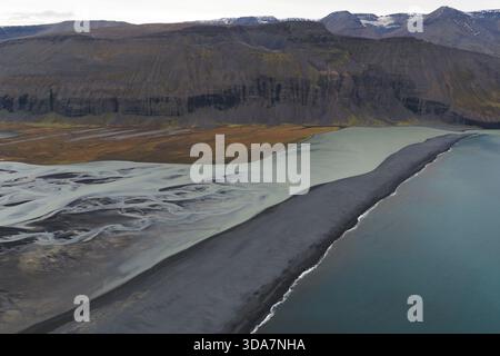Aus der Vogelperspektive auf den starken Kontrast zwischen dem dunklen Strand, dem Gletscherfluss und den zerklüfteten Bergen Island. Stockfoto