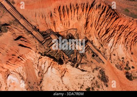 Aus der Vogelperspektive auf die zerklüfteten, sonnengebackenen Klippen und Holztreppen, die hinunter zum Praia dos Tomates führen, wo die Terrakotta-Erde auf den azurblauen Himmel Vilamou trifft Stockfoto