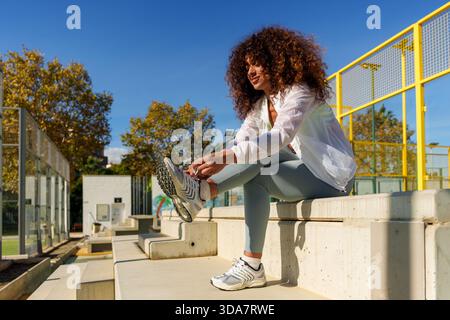 Frau mit lockigem Haar, die ihre Schnürsenkel auf Betonstufen bindet, bereitet sich auf eine Outdoor-Fitness oder Padel-Session vor Stockfoto