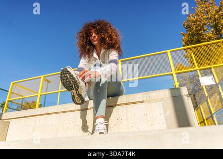 Junge Frau mit lockigen Haaren, die ihren Laufschuh zusammenschnürt, auf Betonstufen sitzt und sich auf eine Fitnessaktivität im Freien vorbereitet Stockfoto