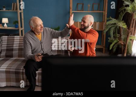Älterer Vater und reifer Sohn geben sich gegenseitig High-Five, während sie zu Hause Videospiele im Fernsehen spielen. Kopierbereich Stockfoto