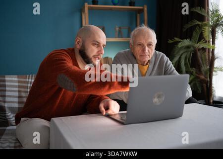 Ein kahlköpfiger erwachsener Sohn hilft dem alten Vater mit einem Laptop, um online zu kommunizieren. Sohn, der Vater im Internet shoppt Stockfoto