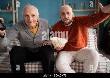 Freizeit für die Familie: Glücklicher Sohn mittleren Alters und älterer Vater, der sich ein aufregendes Fußballspiel auf einem großen Fernsehbildschirm zuhause anschauen, jubeln und essen Stockfoto