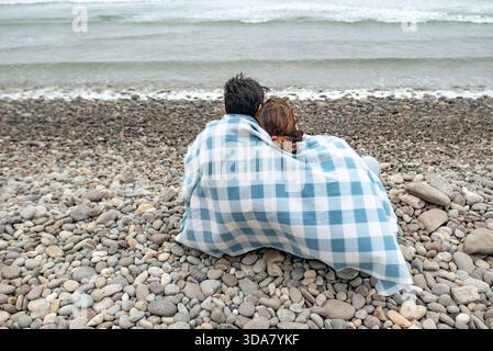 Ein Paar sitzt zusammen unter einer Decke an einem felsigen Strand und blickt auf das Meer und den bewölkten Winterhimmel. Stockfoto