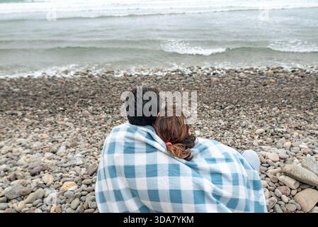 Ein Paar sitzt zusammen unter einer Decke an einem felsigen Strand und blickt auf das Meer und den bewölkten Winterhimmel. Stockfoto