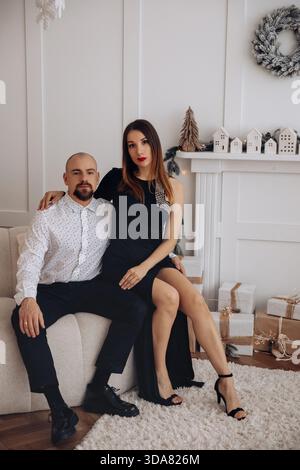 Ein Mann und eine Frau sitzen dicht beieinander auf einer Couch in einem warmen Innenraum. Sie tragen stilvolle Outfits und lächeln, während ein dekorierter Kamin und Gi Stockfoto