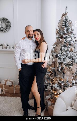 Ein Paar posiert neben einem Weihnachtsbaum in einem festlichen Raum. Die Frau trägt ein schwarzes Kleid, während der Mann Hemd und Hose trägt. Geschenke werden um den herum platziert Stockfoto