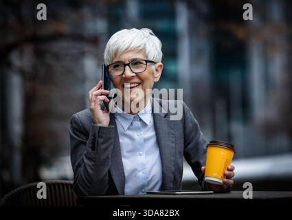 Geschäftsfrau sitzt draußen an einem Tisch, spricht am Telefon und hält eine Kaffeetasse. Sie trägt einen Anzug und hat kurze Haare. Stockfoto