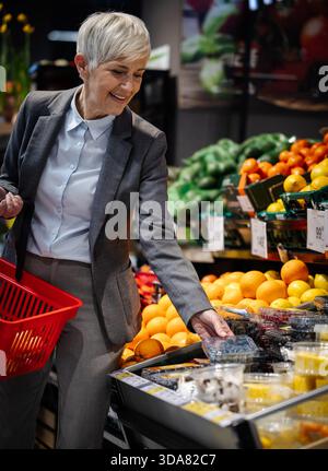 Eine Frau pflückt Blaubeeren in einem Laden. Sie hält einen Korb und lächelt, während sie frische Produkte aus dem Display auswählt. Stockfoto