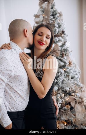 Ein Mann beugt sich vor, um einer Frau auf die Wange zu küssen, während sie neben einem Weihnachtsbaum mit Dekoration steht. Im Winter herrscht eine festliche Atmosphäre Stockfoto