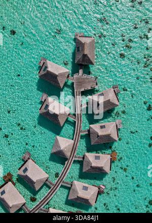 Perfekt vertikale Drohnenaufnahme mit Details zu luxuriösen Bungalows mit Strohdach über dem Wasser, die durch einen gebogenen Holzsteg auf einer klaren türkisfarbenen Lagune verbunden sind. Stockfoto