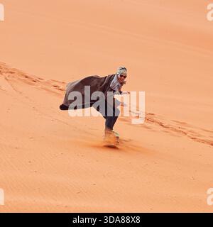 Junger Mann lächelnd, Sanddünen-Surfen im Bist - traditioneller Beduinenmantel. Sandsurfen ist eine der Attraktionen in der Wüste Wadi Rum || Model veröffentlicht Stockfoto