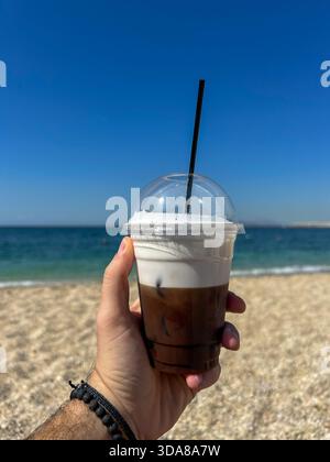 Halten Sie einen Eiskaffee mit Creme vor einem hellblauen Himmel und vor dem Hintergrund des Meeres, um einen erfrischenden Sommermoment am Strand festzuhalten. Stockfoto