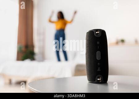Junge Frau tanzt fröhlich in einem Wohnzimmer, während sie Musik von einem kabellosen Lautsprecher auf dem Tisch genießt Stockfoto