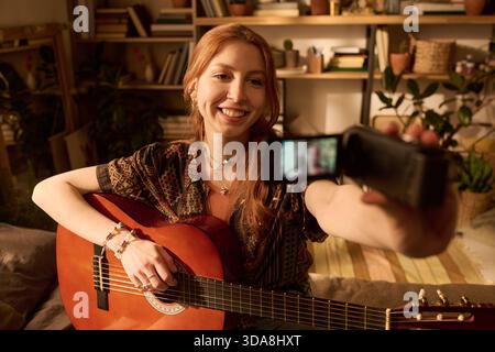 Porträt einer jungen Erwachsenen kaukasischen Frau, die lächelt, während sie Akustikgitarre spielt und sich mit einer tragbaren Videokamera in gemütlicher Atmosphäre aufnimmt, umgeben von Büchern und Pflanzen Stockfoto
