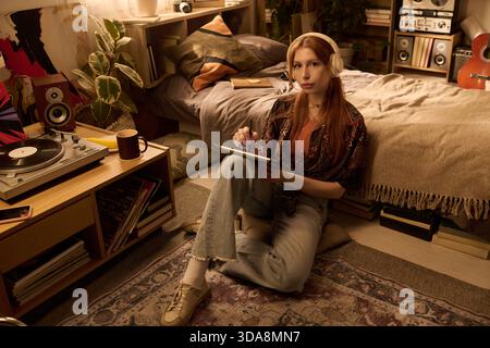Porträt einer kaukasischen jungen erwachsenen Frau, die auf dem Boden in einem gemütlichen Schlafzimmer sitzt und Kopfhörer trägt, die auf einem digitalen Tablet zeichnen, umgeben von Schallplatten und Musikinstrumenten Stockfoto