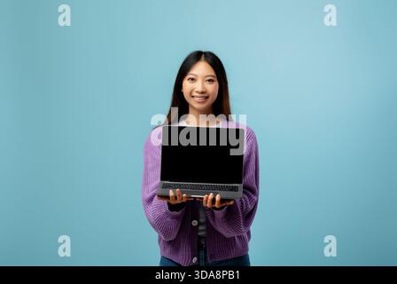 Junge Frau mit Laptop lächelt und steht vor hellblauem Hintergrund in einer ungezwungenen Umgebung Stockfoto