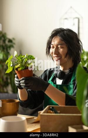 Ein junger asiatischer Blumenhändler lächelt hell, während er vorsichtig eine lebendige grüne Pflanze in den Händen hält. Stockfoto