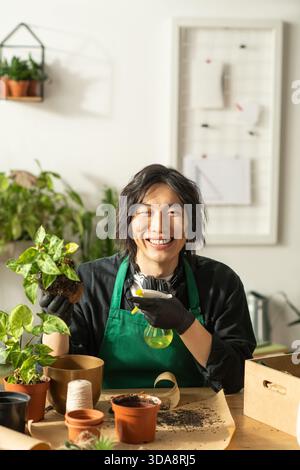 Ein junger asiatischer Mann lächelt hell, während er sich in einer fröhlichen Blumenwerkstatt um Pflanzen kümmert. Stockfoto