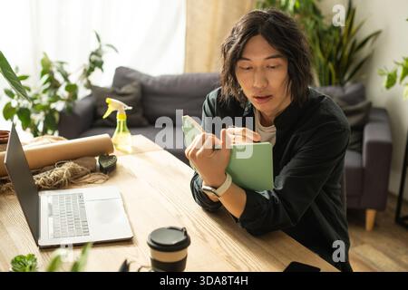 Ein junger asiatischer Blumenhändler skizziert nachdenklich Ideen für Blumendesigns in einem gemütlichen Arbeitsbereich. Stockfoto