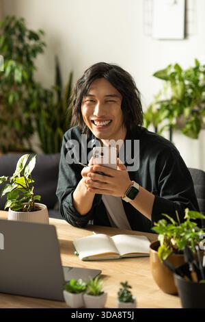 Der junge asiatische Mann lächelt hell, während er sein Smartphone in einer üppigen floralen Umgebung bedient. Stockfoto