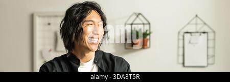 Der junge asiatische Blumenhändler lächelt hell, während er in einer lichtdurchfluteten Werkstatt Blumen arrangiert. Stockfoto
