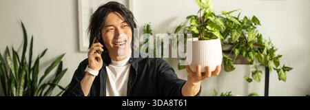 Ein junger asiatischer Blumenhändler lächelt während eines Telefonats hell und zeigt ihm eine Pflanze in der Hand. Stockfoto