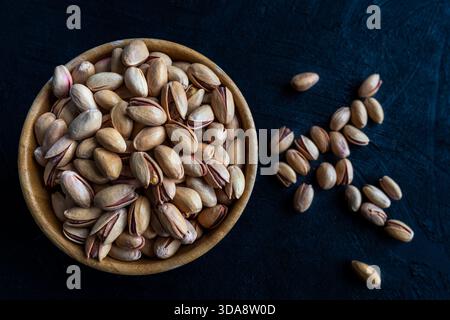 Blick von oben auf eine hölzerne Schüssel gefüllt mit gerösteten Pistazien, mit einigen Nüssen, die auf einer strukturierten dunklen Oberfläche verstreut sind. Stockfoto