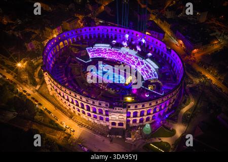 Aus der Vogelperspektive auf die Pula Arena, in pulsierende lila und blaue Lichter getaucht, um Schlittschuhlaufen zu können, was ein faszinierendes Spektakel schafft, Pula, Kroatien. Stockfoto