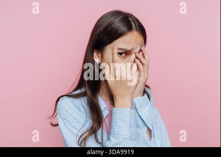 Frau, die durch die Finger blickt, mit einem besorgten Ausdruck vor einem rosafarbenen Hintergrund. Stockfoto