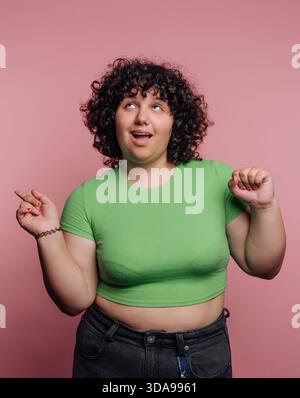 Eine fröhliche Frau mit lockigem Haar, die ein grünes Oberteil trägt, posiert spielerisch vor einem rosa Hintergrund. Stockfoto