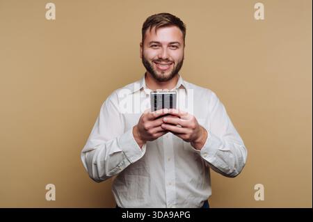 Lächelnder Mann in weißem Hemd, der ein Smartphone vor beigefarbenem Hintergrund hält. Stockfoto