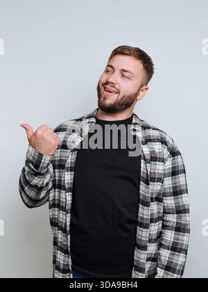 Mann in kariertem Hemd, der mit Daumen zeigt, vor grauem Hintergrund lächelnd. Stockfoto